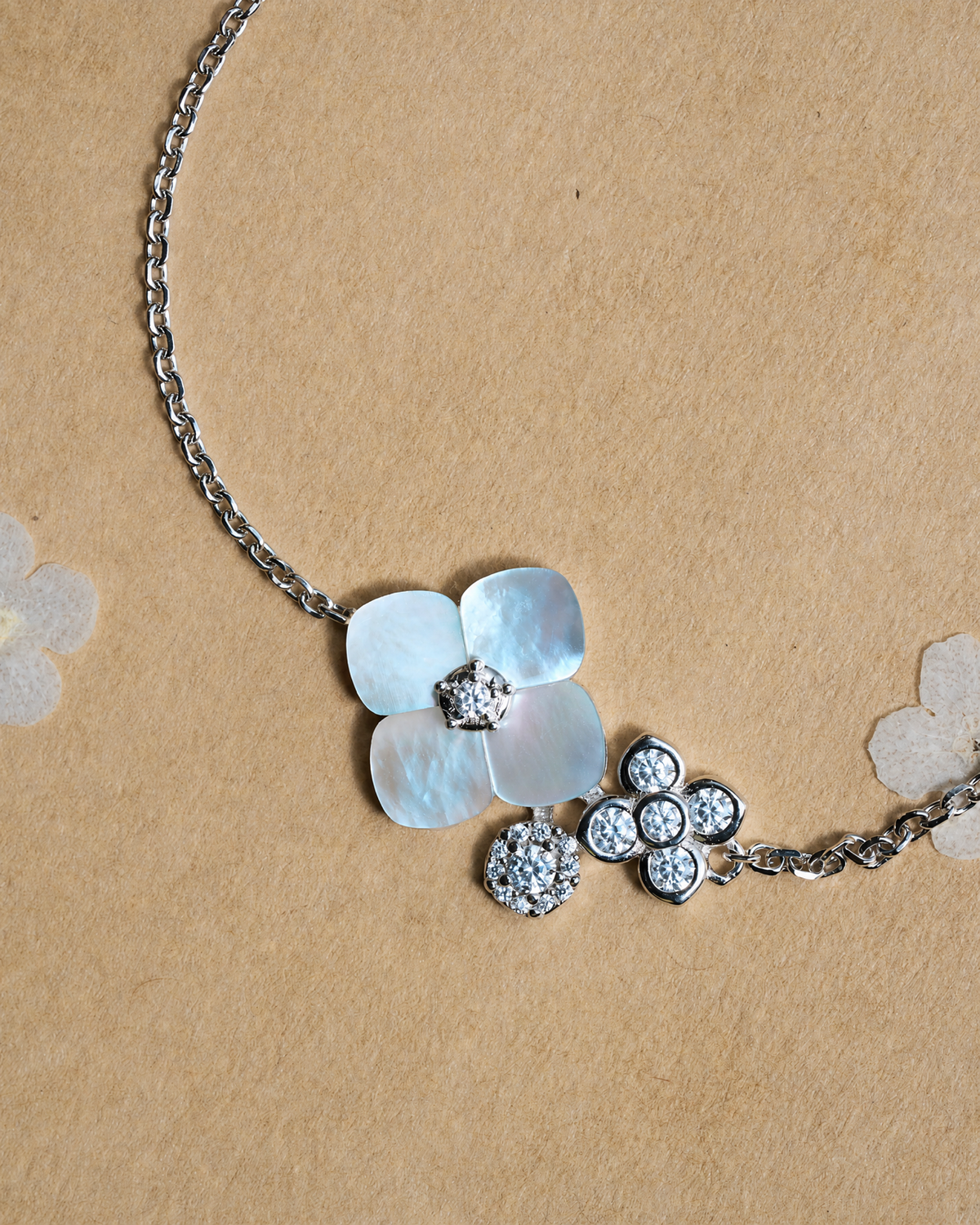 A silver-tone floral necklace featuring a large mother-of-pearl flower with two small zircon flowers on the side, accompanied by smaller crystal-accented flowers and a chain, displayed on a textured tan paper background.