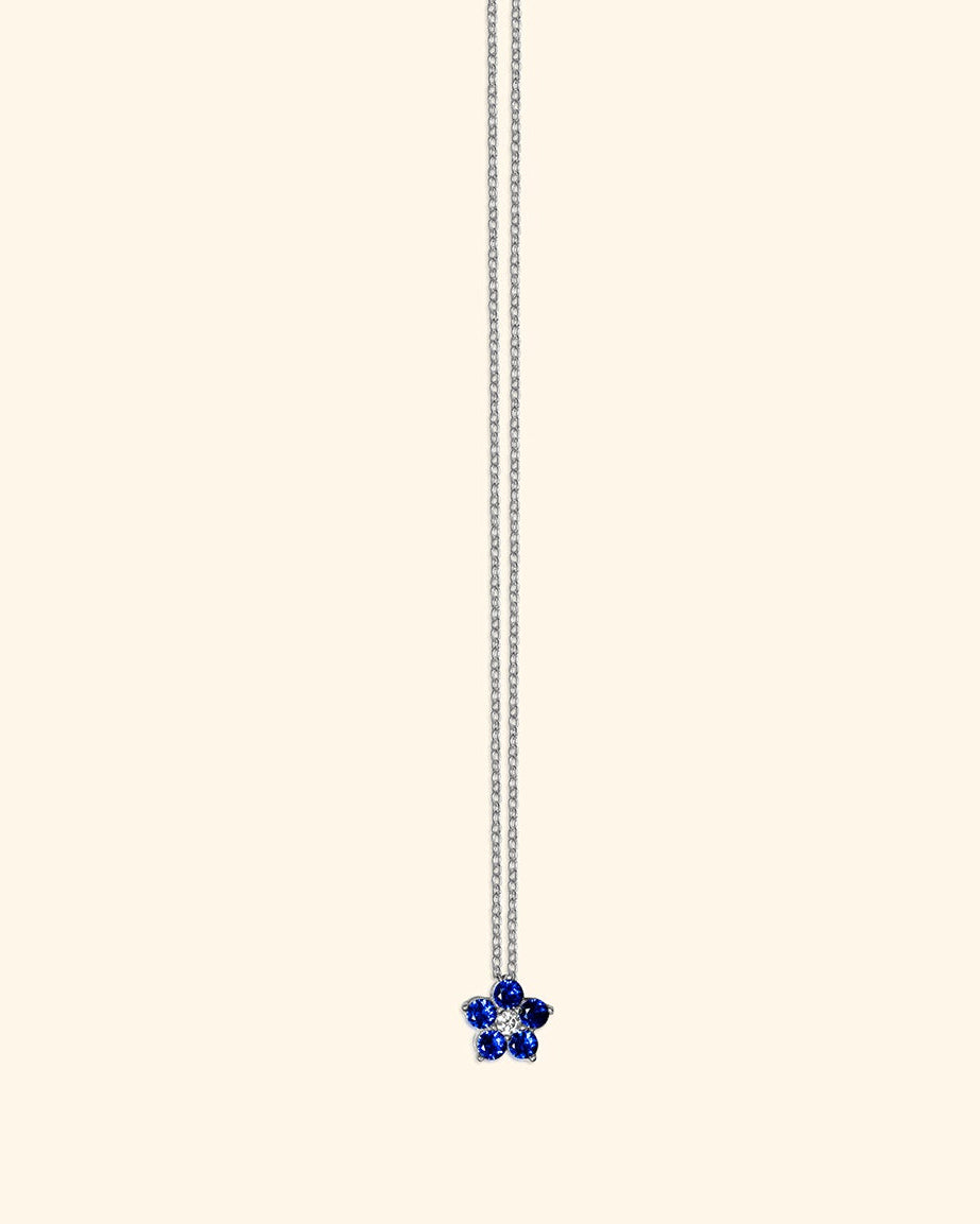 A delicate silver chain necklace featuring a small, five-petal flower charm set with deep blue stones at the bottom, presented on a solid cream background.