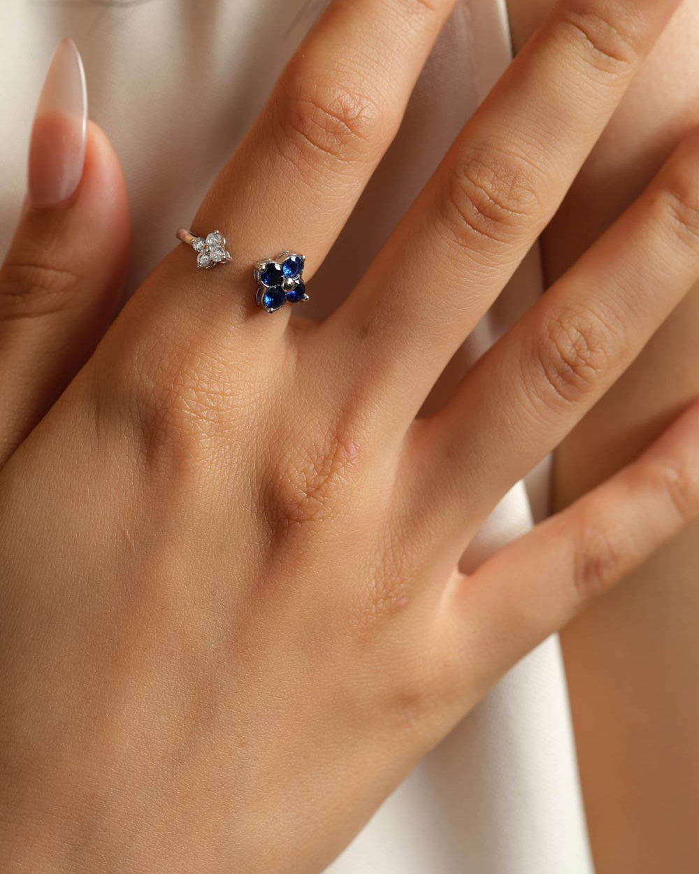 A close-up of a hand wearing two small rings: one silver-toned ring with a white flower zircon and one silver-toned ring with a blue flower-shaped zircon, against a soft cream background.