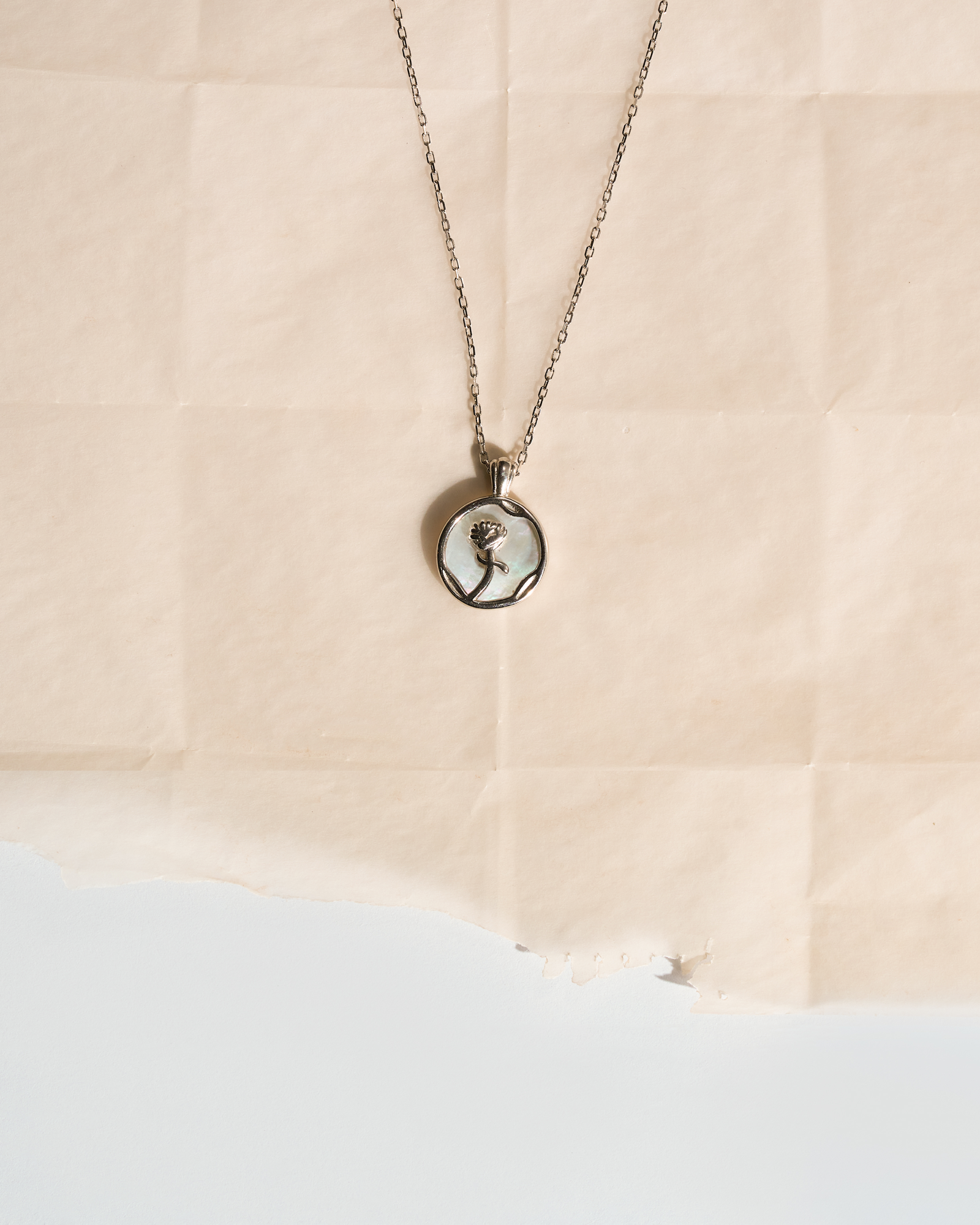 A silver-toned chain necklace with a round pendant featuring a black-and-white portrait of a woman, displayed on a torn sheet of beige paper against a white background.