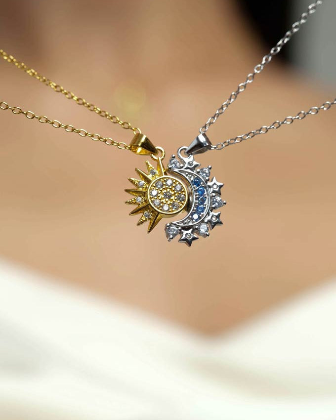 A close-up of two delicate necklaces worn against a blurred skin background: one gold chain with a sun pendant set with yellow stones, and one silver chain with a crescent moon and star pendant accented by blue stones.