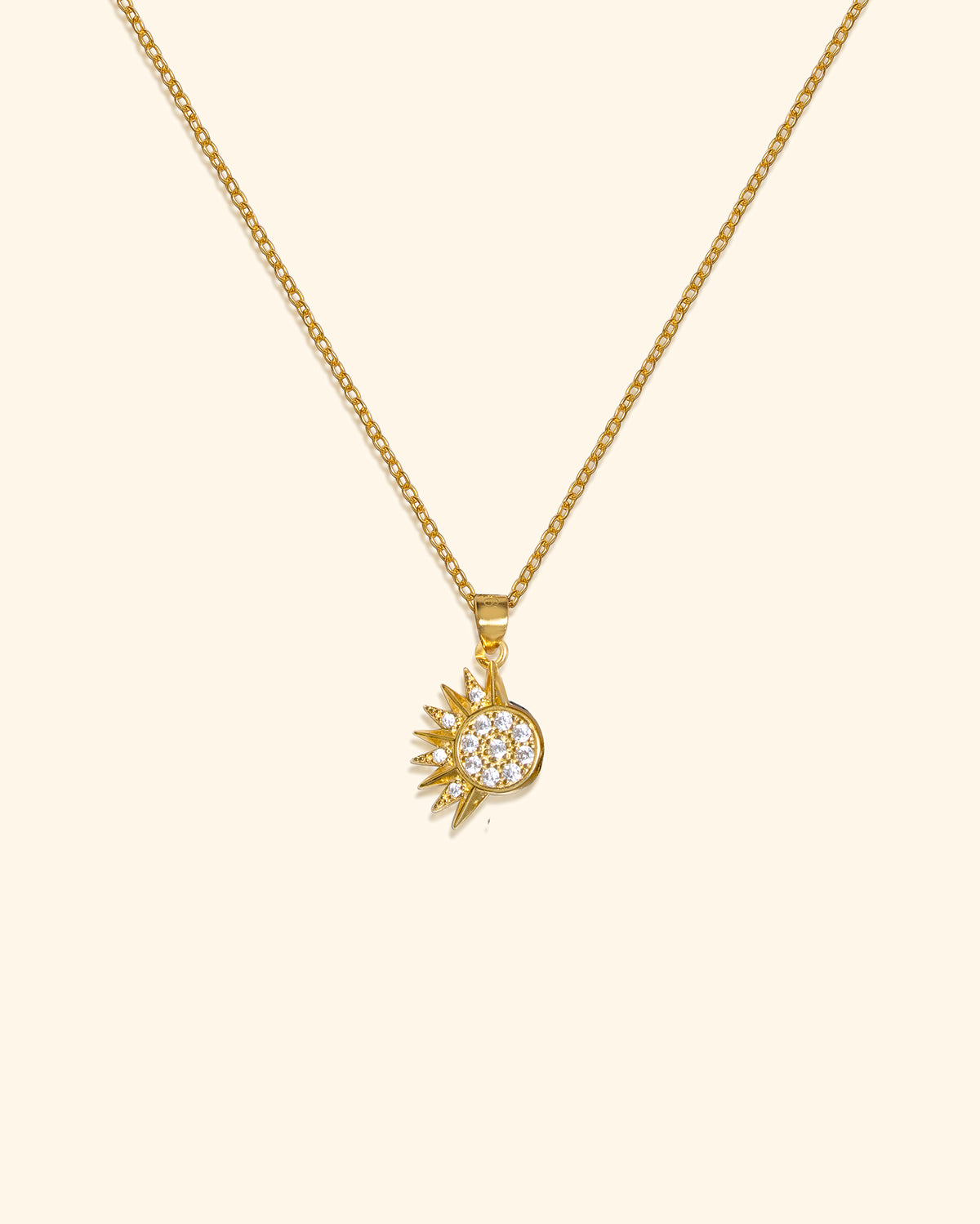 A gold chain necklace with a small sunburst pendant featuring a circular cluster of clear pavé stones, shown on a plain cream background.