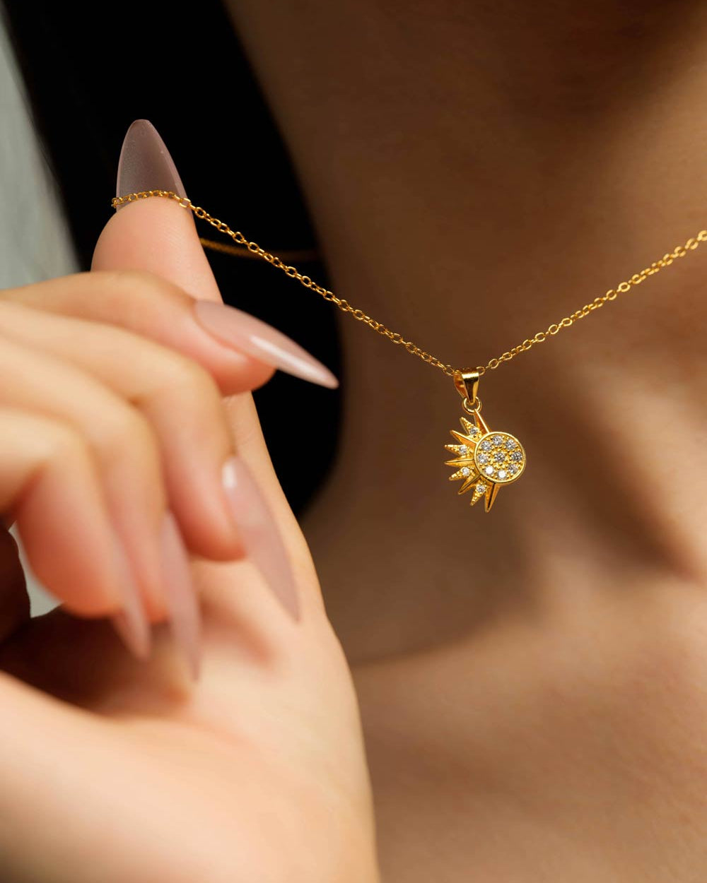 A gold necklace with a sun pendant set with small clear stones, worn on a model's neck and held by a hand with long nude manicured nails.