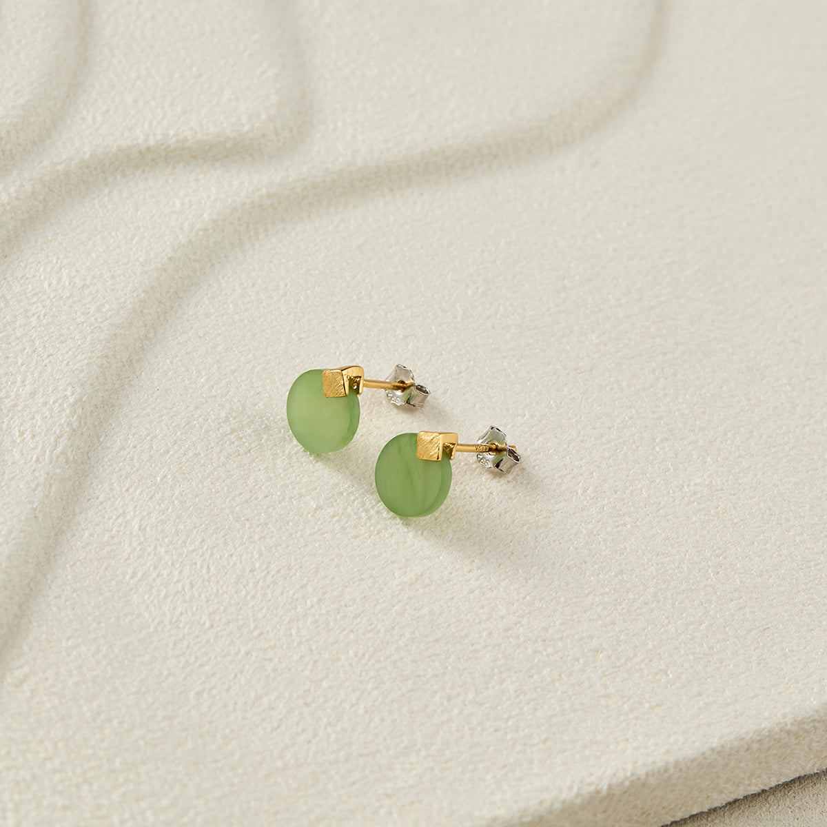 Still life photography of Choodo natural green Jade sterling silver earrings laid flat on a serene beige tabletop, modern intellectual premium vibe.
